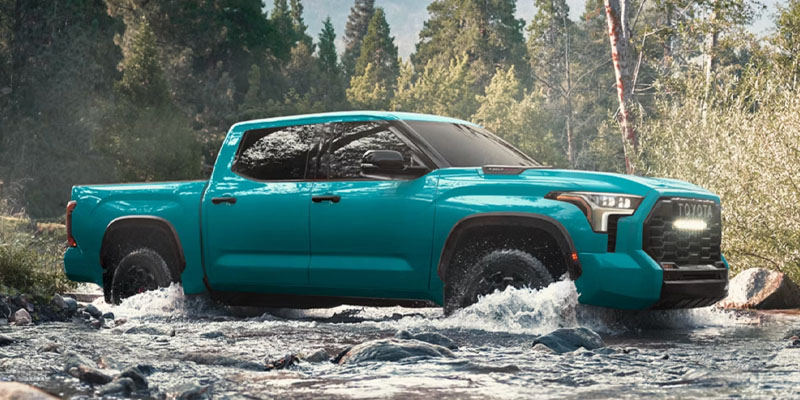 Teal Toyota Tundra pickup truck splashing through a river in a forest.