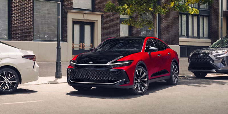 Red and black modern crossover SUV parked on a city street, flanked by other vehicles with brick buildings and trees in the background.