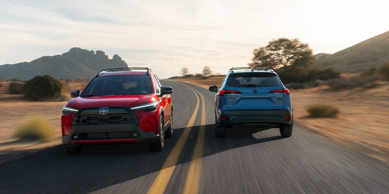 Two Toyota Toyota Corolla Cross SUVs driving side by side on a scenic open road with mountains in the background.